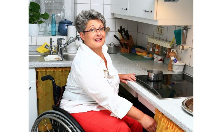 Eine Frau mit kurzen, grauen Haaren und Brille sitzt in einem Rollstuhl vor der Kochplatte in ihrer Küche.o