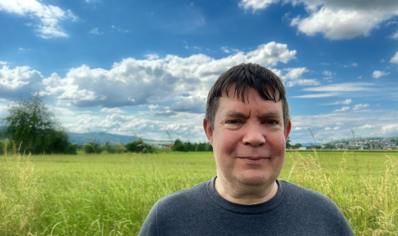 Ein Mann blickt in die Kamera. Er steht draussen in der Natur. Im Hintergrund eine grüne wiese und ein tiefblauer Himmel mit weissen Wolken.