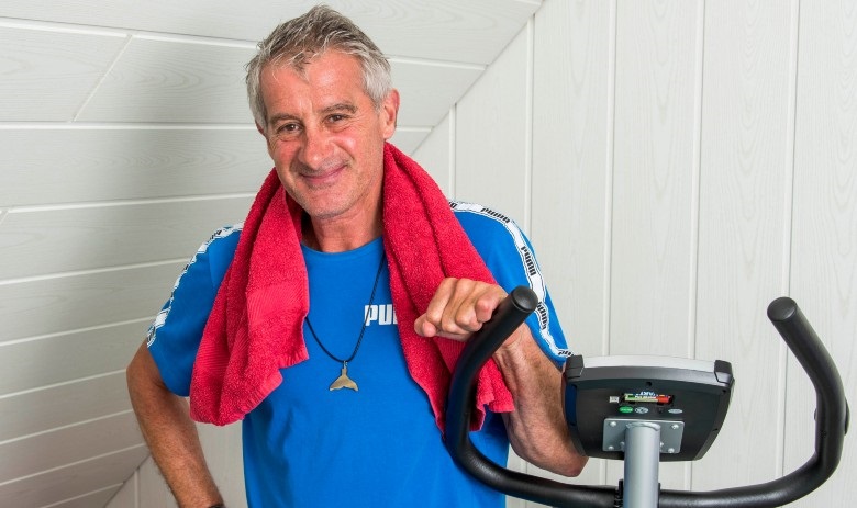 Daniel S. Portrait eines Mannes, der neben seinem Home-Trainer steht. Er trägt ein Blaues Sport-Shirt und hat ein rotes Tuch um den Nacken.